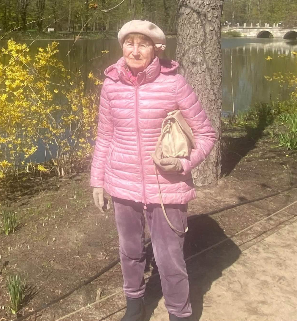 Zdjęcie przedstawia starszą kobietę ubraną w różową kurtkę z beretem na głowie stojącą na dworze.