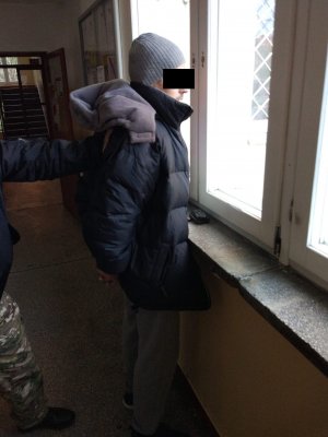 Na zdjęciu widać rękę policjanta przytrzymującą za bark zatrzymanego mężczyznę. Zatrzymany mężczyzna stoi przodem do okna.