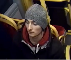 Na zdjęciu widać mężczyznę zajmującego miejsce siedzące w autobusie frontem do kamery. Mężczyzna ma na sobie szarą czapkę, czarną kurtkę spod której wystaje bluza z kapturem koloru bordowego. W tle widać wolne siedzenia dla pasażerów.