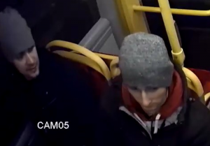 Na zdjęciu widać dwóch mężczyzn zajmujących miejsca siedzące w autobusie frontem do kamery. Obaj ubrani są w szare czapki.