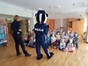 Na zdjęciu widać stojące tyłem policjantki, jedna w stroju maskotki Borsuka. W tle widać siedzące na ławeczkach dzieci.
