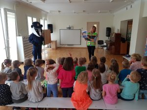 Na zdjęciu widać siedzące tyłem dzieci, policjantkę przebraną w strój Borsuka i kobietę ubraną w żółtą kamizelkę odblaskową, która pokazuje dzieciom książeczkę