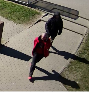 Na zdjęciu widać dwóch mężczyzn, którzy przebywają na zewnątrz. Jeden ubrany jest na ciemno, ma założony kaptur, drugi ma czerwoną kurtkę, czerwone buty, ciemne spodnie