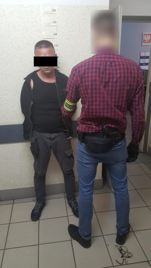 Zdjęcie przedstawia policjanta ubranego po cywilnemu, który ma na lewym przedramieniu opaskę z napisem &quot;POLICJA&quot;. Policjant przytrzymuje stojącego pod ścianą zatrzymanego mężczyznę.