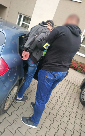 Na zdjęciu widać mężczyznę w kajdankach, podtrzymywanego przez policjanta, który pomaga mu wsiąść do radiowozu