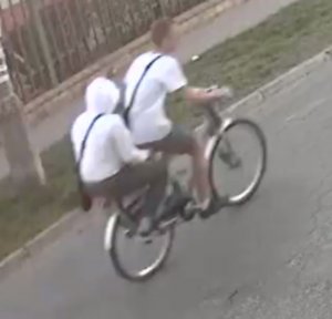 Zdjęcie przedstawia dwie osoby jadące rowerem, jedna siedzi na bagażniku. Osoby ubrane są w białą koszulkę i białą bluzę. Mają przewieszone przez klatkę piersiową czarne torebki