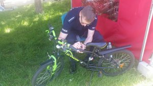 Zdjęcie przedstawia policjanta ubranego w mundur, który pochyla się na rowerem i specjalnym narzędziem wykonuje grawer na ramie. W tle widać ścianę namiotu.
