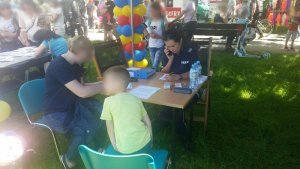 Na zdjęciu widać policjantkę, która siedzi przy stole razem z małym chłopcem i kobietą i wypełnia leżące przed nią dokumenty. Na około widać zgromadzonych przy innych stoiskach ludzi.