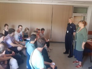 Na zdjęciu widać grupę osób siedzących na sali. Przed nimi stoi policjantka ubrana w granatowy mundur i kobieta ubrana w zieloną bluzkę i niebieską spódnicę. Obie są skierowane frontem do siedzących osób.