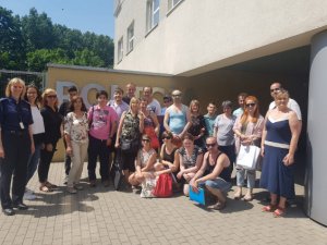 Zdjęcie przedstawia grupę osób stojących przed budynkiem komendy i uśmiechających się do zdjęcia. Za nimi znajduje się ściana i napis "POLICJA".