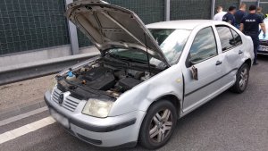 Zdjęcie przedstawia srebrny pojazd osobowy marki Volkswagen, pojazd ma uniesioną przednią klapę. Zaparkowany jest na pasie ruchu, w tle widać osłony dźwiękoszczelne trasy S8. Samochód posada liczne wgniecenia na lewym boku, ma też urwane lusterko boczne. Za nim widać grupę stojących tyłem policjantów w granatowych mundurach i ubranych po cywilnemu.
