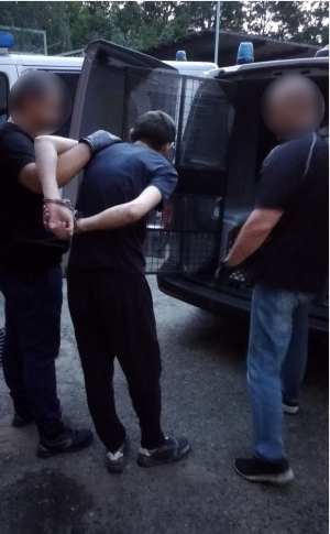 Zdjęcie przedstawia policjanta w ubraniu cywilnym stojącego tyłem i trzymającego za ręce mężczyznę, który ma założone kajdanki na ręce trzymane z tyłu. Zatrzymany mężczyzna jest pochylony do przodu. Przed nimi znajduje się radiowóz z otwartymi zakratowanymi drzwiami. Po prawej stronie zatrzymanego mężczyzny znajduje się drugi policjant w ubraniu cywilnym, ma założone na dłonie ciemne rękawiczki.