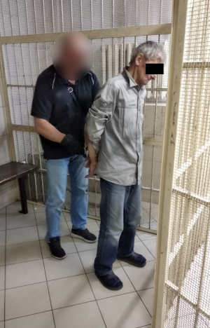 Zdjęcie przedstawia policjanta, który zakłada kajdanki mężczyźnie stojącemu do niego tyłem i trzymającego ręce z tyłu. W tle widać kraty pomieszczenia dla osób zatrzymanych