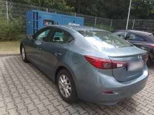 Zdjęcie przedstawia pojazd osobowy marki Mazda w kolorze srebrnym. Pojazd zaparkowany jest przed niebieską skrzynką. Po prawej stronie widać inny zaparkowany równolegle samochód osobowy.