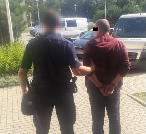Na zdjęciu widać mężczyznę w kajdankach, z rękami z tyłu, którego prowadzi policjant. W tle widać zaparkowane cywilne samochody