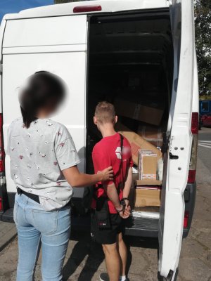 Zdjęcie przedstawia policjantkę w ubraniu cywilnym, która trzyma za lewe przedramię mężczyznę z założonymi kajdankami na ręce trzymane z tyłu. Mężczyzna ma czerwoną koszulkę na krótki rękaw z białym napisem na plecach, czarne krótkie spodenki, czarne buty sportowe i białe skarpetki oraz czarną saszetkę przewieszoną przez prawe ramię. Obie osoby stoją przodem do zaparkowanego białego dostawczego fiata. Pojazd ma otwarte tylne drzwi od część bagażowej. Wewnątrz widać brązowe pudełka.