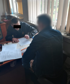 Zdjęcie przedstawia siedzącego przy biurku w pokoju służbowym policjanta w cywilnym ubraniu, który wykonuje czynności z zatrzymanym mężczyzną. Mężczyzna ten siedzi przy tym samym biurku naprzeciwko policjanta i podpisuje dokument leżący na biurku. Na blacie biurka rozłożone są inne dokumenty. Z prawej strony widać zasłonięte okno.