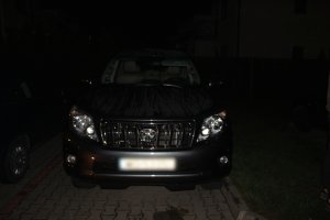 Zdjęcie przedstawia stojący przodem samochód osobowy. Pojazd jest ciemnego koloru, ma zasłoniętą przednią tablicę rejestracyjną. Samochód jest fotografowany nocą. Po lewej stronie zdjęcia widać fragment innego zaparkowanego samochodu. Z tyłu widać biały budynek mieszkalny i ogrodzenie.