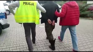 Zdjęcie przedstawia dwóch policjantów w ubraniu cywilnym prowadzących zatrzymanego mężczyznę po parkingu. Mężczyzna ma założone kajdanki na ręce trzymane z tyłu. Policjant idący z lewej strony mężczyzny ma założoną żółtą kamizelkę z napisem POLICJA. Z lewej strony zdjęcia widać fragmenty radiowozów policyjnych.