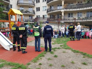 Zdjęcie przedstawia dzieci ustawione w szeregu na dworze, którym towarzyszą opiekunki. Przed nimi stoją strażacy, policjantka, przedstawiciel urzędu dzielnicy w odblaskowej kamizelce. W tle widać bloki mieszkalne. Po lewej stronie zdjęcia widać elementy placu zabaw.