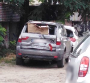 Zdjęcie przedstawia pojazd m-ki BMW. Ma on uszkodzoną tylną szybę zasłoniętą brązowym kartonem. Przed nim zaparkowany jest inny pojazd koloru białego. W tle widać fragment budynku.