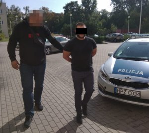 Zdjęcie przedstawia policjanta ubranego w cywilną odzież prowadzącego zatrzymanego mężczyznę. Mężczyzna ten trzyma ręce z tyłu, ma założone kajdanki. Ubrany jest w szare długie spodnie i czarną koszulkę. Obok mężczyzny (z prawej strony zdjęcia) widać fragment oznakowanego radiowozu. W tle widać inne zaparkowane pojazdy.