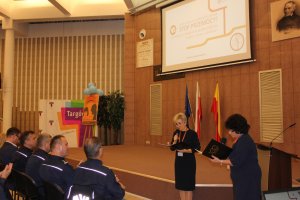 Zdjęcie przedstawia wnętrze sali w urzędzie. Na zdjęciu widać dwie kobiety ubrane w ciemne sukienki, które trzymają w rękach rozłożone teczki i mikrofony. W tle, na ścianie widać wyświetlony slajd z napisem &quot;STOP PRZEMOCY&quot;. Pod spodem znajdują się trzy kolorowe flagi. Z lewej strony zdjęcia widać plakat z napisem Targówek oraz kolorowy plakat umieszczony na scenie. W pierwszym rzędzie przed sceną (tyłem do aparatu) siedzą policjanci w granatowym służbowym mundurze.