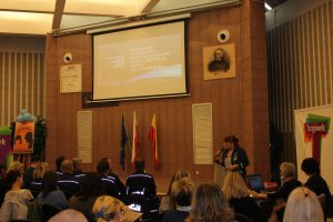 Zdjęcie przedstawia wnętrze sali z uczestnikami konferencji. Goście siedzą na krzesłach tyłem do aparatu. Wpatrują się w kobietę, która stoi po prawej stronie zdjęcia i trzyma mikrofon. Na wprost widzów wyświetlony jest slajd o treści: &quot;Zrozumieć nastolatka - samookaleczenia, apatia, depresja...moda czy choroba?&quot;