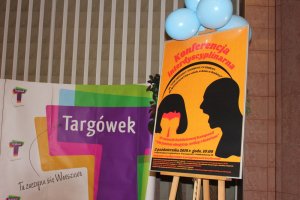 Zdjęcie przedstawia kolorowy plakat pod hasłem: &quot;Konferencja interdyscyplinarna&quot;. Plakat umieszczony jest na brązowym drewnianym stelażu. Nad plakatem znajdują się niebieskie balony. W tle, po lewej stronie zdjęcia widać inny kolorowy plakat z napisem &quot;Targówek. Tu zaczyna się Warszawa&quot;.