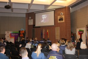 Zdjęcie przedstawia uczestników konferencji siedzących na krzesłach (tyłem do aparatu). Widzowie wpatrują się w slajd umieszczony na wyświetlaczu przed nimi. Z prawej strony widzów widać kobietę, która również wpatruje się w slajd.