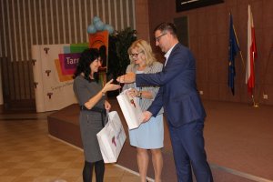 Zdjęcie przedstawia kobietę o blond włosach, w okularach o czarnej oprawce, ubraną w biało-szarą marynarkę i niebieską sukienkę i mężczyznę ubranego w ciemny granatowy garnitur i białą koszulę, którzy wręczają kobiecie ubranej w szarą sukienkę torebkę z logiem: &quot;Targówek&quot;. Kobieta uśmiecha się, w jednej ręce trzyma już jedną taką torbę.