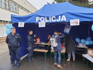 Zdjęcie przedstawia niebieski namiot z napisem na górze POLICJA. W środku widać dwie policjantki w granatowych mundurach. Jedna z nich siedzi za stołem. Po lewej stronie zdjęcia widać stojącego policjanta-dzielnicowego w granatowym mundurze. Wewnątrz namiotu znajdują się ławki i stoły, na których leżą różne materiały. W tle widać budynek z szarą elewacją.
