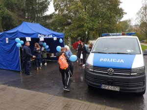 Zdjęcie przedstawia niebieski namiot z napisem POLICJA,który znajduje się na otwartej przestrzeni. Jest ozdobiony niebieskimi balonami. Widać przy nim kręcących się ludzi. Po prawej stronie stoi policjant w granatowym mundurze. Na zdjęciu jest też oznakowany radiowóz Policji.