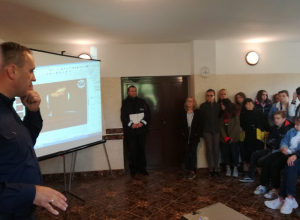Zdjęcie przedstawia kilkudziesięciu uczniów siedzących na krzesłach w pomieszczeniu. Wpatrują się one w tablicę na której wyświetlony jest slajd. Z lewej strony zdjęcia widać policjanta w granatowym mundurze służbowym oraz policjanta stojącego w drzwiach.
