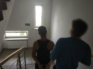 Zdjęcie przedstawia klatkę schodową i policjanta w umundurowaniu cywilnym, który prowadzi zatrzymanego mężczyznę. Ma on kajdanki na rękach trzymanych z tyłu. Mężczyzna ubrany jest w ciemną koszulkę tylu &quot;bokserka&quot;. Z lewej strony zdjęcia widać poręcz schodów.
