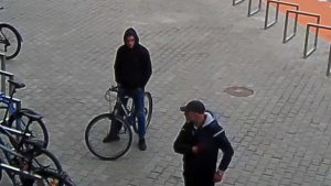 Zdjęcie przedstawia mężczyznę w wieku około 25-30 lat, wzrost 175-180 cm, ubranego w czarną bluzę z kapturem, spodnie jeansowe niebieskie, czarne buty sportowe, na głowie ma założony kaptur, który stoi z rowerem i trzyma ręce w kieszeniach spodni. Obok niego znajduje się drugi mężczyzna w wieku około 25-30 lat, wzrost około 170-175 cm, ubrany w granatową bluzę z kapturem, na głowie ma założoną czarną czapkę z daszkiem, który prawą ręką sięga pod materiał bluzy. Z lewej strony zdjęcia widać zaparkowane przy stojaku rowery.