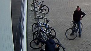 Zdjęcie przedstawia mężczyznę w wieku około 25-30 lat, wzrost 175-180 cm, ubranego w czarną bluzę z kapturem, spodnie jeansowe niebieskie, czarne buty sportowe, który stoi z rowerem i przygląda się drugiemu mężczyźnie ubranemu w granatową bluzę z kapturem, który kuca przy jednym z stojących przy stojaku rowerze. W tle widać inne rowery pozostawione przy stojaku.