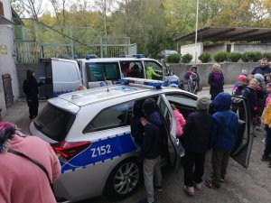 Zdjęcie przedstawia dziedziniec komisariatu, na którym zaparkowane są dwa oznakowane radiowozy. Pojazdy mają otwarte drzwi, do środka wchodzą uczniowie. Część osób stoi obok zaparkowanych samochodów. Przy większym radiowozie, z tyłu stoi policjantka w granatowym mundurze, która trzyma drzwi.
