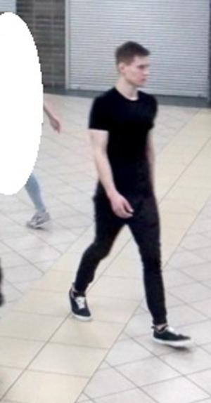Zdjęcie przedstawia dwóch mężczyzn poruszających się w obiekcie dworca. Pierwszy mężczyzna w wieku około 25 lat, normalnej sylwetki, średniego wzrostu, ma czarne krótkie włosy, okulary korekcyjne, ubrany jest w czerwoną bluzę z kapturem, czarne krótkie spodenki, czarne buty, na ramieniu ma plecak koloru czarnego. Prowadzi czarnego dużego psa – prawdopodobnie doga, którego trzymał na czerwonych szelkach i smyczy. Z jego lewej strony idzie mężczyzna w wieku około 25 lat, szczupłej sylwetki, wysoki, ma włosy ciemny blond, ubrany jest w czarny t-shirt, czarne spodnie i czarne buty.