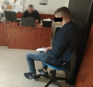 Zdjęcie przedstawia policjanta w ubraniu cywilnym, który siedzi w pomieszczeniu służbowym, przy biurku. Przed nim stoi czarny laptop, drugi laptop znajduje się po jego prawej stronie. Przed biurkiem na niebiesko-czarnym krześle siedzi zatrzymany mężczyzna. Ubrany jest w granatową kurtkę, niebieskie spodnie jeansowe i czarne buty sportowe. W rękach trzyma kartkę. Wpatruje się w podłogę. Za policjantem, na ścianie wisi tablica, a na niej umieszczone kartki.
