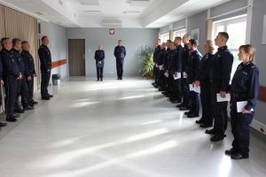 Zdjęcie przedstawia stojących w sali policjantów w granatowych mundurach służbowych. Z prawej strony stoją w rzędzie policjanci, którzy otrzymali awanse. Naprzeciwko nich stoi kadra kierownicza jednostki.