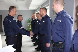 Zdjęcie przedstawia stojących w rzędzie policjantów w granatowych mundurach służbowych. Naprzeciwko jednego z nich stoi Komendant, który podaje mu rękę, obok widać Zastępcę Komendanta, który podaje rękę innemu policjantowi.
