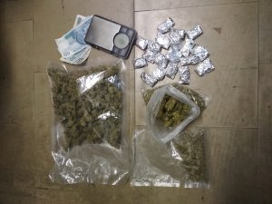 Zdjęcie przedstawia leżące na ciemnym podłożu zabezpieczone przedmioty. Wśród nich jest telefon komórkowy leżący wyświetlaczem do dołu, ma szaro-czarną obudowę; częściowo pod nim leżą trzy banknoty: dwa o nominale 50 złotych i jeden - 100 złotych; trzy foliowe torebki z suszem roślinnym oraz 19 zawiniątek ze srebrnej folii.