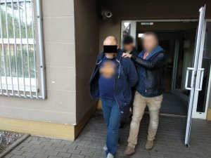Zdjęcie przedstawia policjanta w cywilnych ubraniach prowadzącego zatrzymanego mężczyznę. Mężczyzna trzyma ręce z tyłu. Ubrany jest w granatową rozpiętą bluzę, granatową koszulkę z pomarańczowym nadrukiem, granatowe spodnie jeansowe i szare buty. Policjant idzie z jego lewej strony, jedną ręką przytrzymuje go za ramię. Z tyłu widać fragment drugiego policjanta. Wszyscy wychodzą z budynku, drzwi wyjściowe są szeroko otwarte. Po prawej stronie mężczyzn widać zakratowane okno pomieszczenia dla osób zatrzymanych.