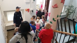 Zdjęcie przedstawia schodzące po schodach w obiekcie dzieci. Za nimi widać dwie opiekunki. Po ich lewej stronie stoi policjantka w granatowym mundurze.