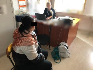 Zdjęcie przedstawia pokój służbowy policjantów, przy biurku przed otwartym laptopem siedzi ubrana w cywilne ubrania policjantka. Przed nią na krześle, siedzi zatrzymana kobieta. Ma ona ciemne włosy, kolorową kurtkę i ciemne spodnie. Na podłodze, oparty o biurko leży jasny plecak i zielona torba.