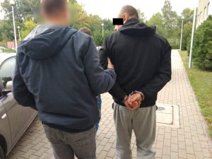 Zdjęcie przedstawia policjanta w ubraniu cywilnym, który prowadzi zatrzymanego mężczyznę. Mężczyzna ma kajdanki założone na ręce trzymane z tyłu. Ubrany jest w czarną kurtkę z kapturem oraz jasnoszare spodnie materiałowe w kratkę. Z lewej strony zdjęcia widać fragment pojazdu osobowego w ciemnym kolorze; z prawej - ścianę budynku, latarnie i pas zieleni.