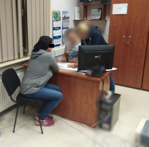Zdjęcie przedstawia pokój służbowy policjantów. Przy biurku, na którym stoi monitor laptopa, bokiem do aparatu siedzi zatrzymana przez policjantów kobieta. Ma ona czarne rozpuszczone włosy. Ubrana jest w szarą bluzę, niebieskie spodnie jeansowe i fioletowe adidasy. Naprzeciwko niej jest dwoje policjantów, którzy wpatrują się w dokumenty rozłożone na blacie biurka. Oboje są w ubraniach cywilnych. Na ścianie widać kalendarz, za policjantami znajduje się brązowa szafa, na której wisi biała kartka. Biurko stoi przy oknie.