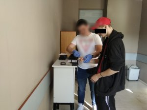 Zdjęcie przedstawia pokój służbowy. Przy wysokim jasnym stoliku, ustawionym pod ścianą, stoi policjant w ubraniu cywilnym, który ma założone niebieskie rękawiczki ochronne. Policjant trzyma prawą dłoń zatrzymanego mężczyzny, nad białą kartką. Mężczyzna ma na sobie czerwoną czapkę z daszkiem, czarną rozpiętą bluzę, pod którą widać czerwoną koszulkę i czarne spodnie z białymi paskami. W tle, w lewym rogu pomieszczenia widać szafę drewnianą koloru brązowego, w prawym rogu ustawiona jest pod ścianą niszczarka.