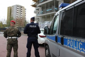 Zdjęcie przedstawia policjanta w granatowym mundurze stojącego obok żandarma. Z prawej strony funkcjonariuszy stoi policyjny radiowóz. W tle widać bloki mieszkalne